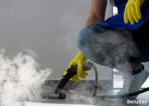 A person wearing yellow rubber gloves and a blue apron uses a steam cleaner to remove mold from a surface, with thick steam visibly rising. The "BenzVac" branding is in the bottom right corner. How Long Does Mold Remediation Take